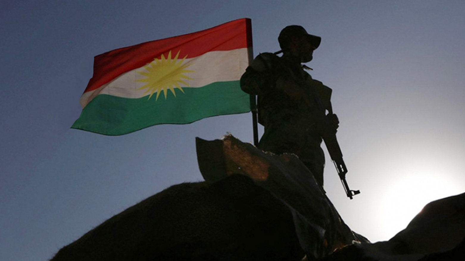 A silhouette of a Peshmerga member standing beside the Kurdistan flag, which is prominently waving against a clear sky. (Photo: Kurdistan24)