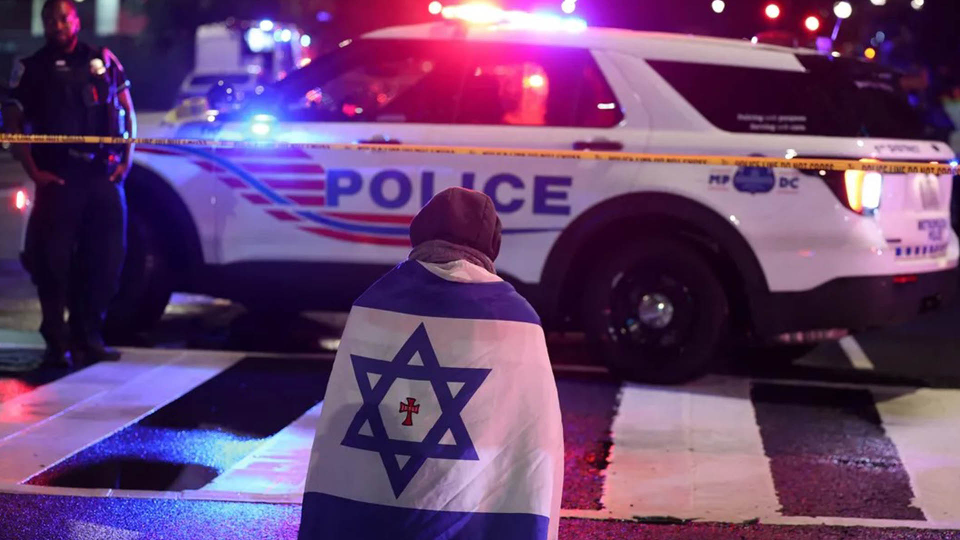 man draped in an Israeli flag with a cross on it gestures as police secure the area outside the Capital Jewish Museum after a shooting left two dead in Washington, DC, on May 22, 2025. (Photo: AFP)