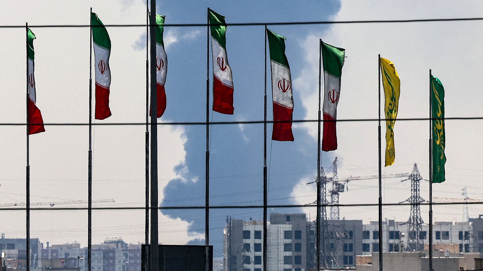 Flags flutter along a bridge as a plume of heavy smoke and fire rise from an oil refinery in southern Tehran, after it was hit in an overnight Israeli strike, on Jun.15, 2025. (Photo: AFP)