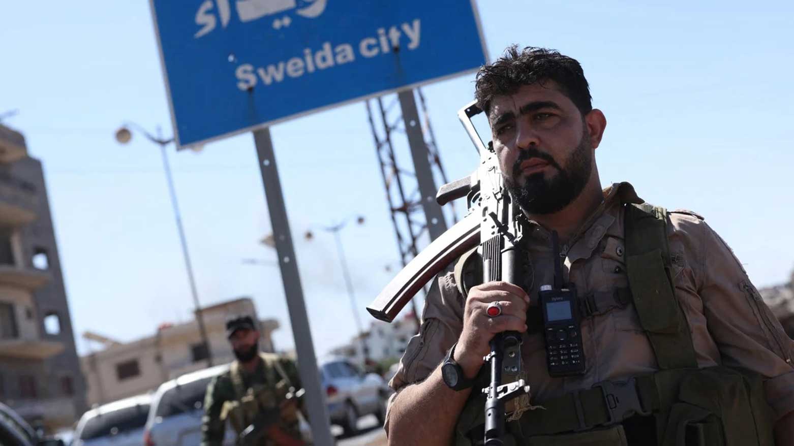 A member of Syria's security forces stands under a sign in the predominantly Druze city of Sweida, 15 July (Photo: AFP)