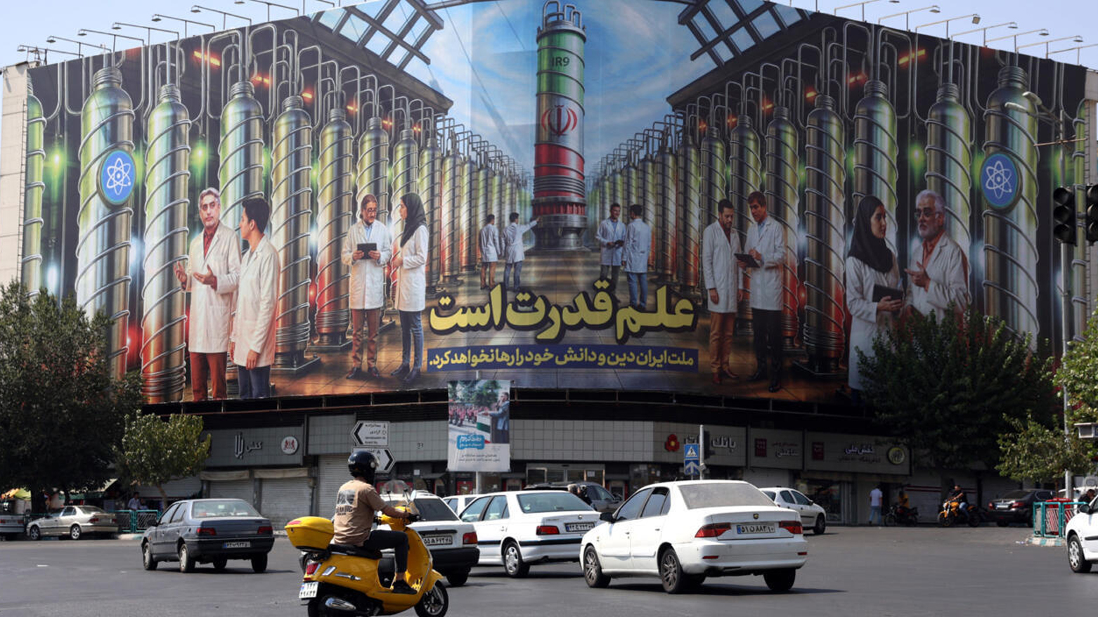 ایرانیان در کنار بیلبوردی که تصاویر دانشمندان هسته‌ای، سانتریفیوژها و جمله‌ای به فارسی با مضمون «علم قدرت است» را نشان می‌دهد – عکس: ای‌اف‌پی