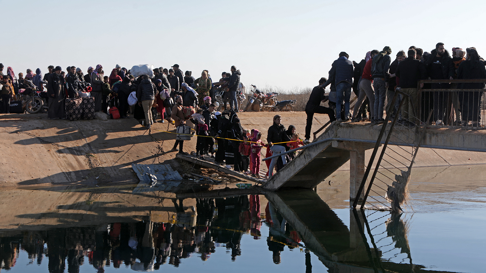 People along with their belongings walk across a damaged bridge as they flee from a Kurdish-controlled area, in Rasm al-Harmal, east of Aleppo city on January 15, 2026. (AFP)