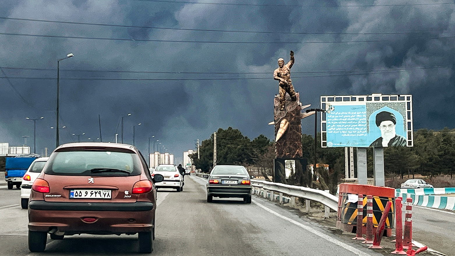 Vehicles move along a highway past a war memorial statue and a billboard depicting Iran's late supreme leader Ayatollah Ali Khamenei, with plumes of black smoke billowing, in Tehran on March 8, 2026. (AFP)