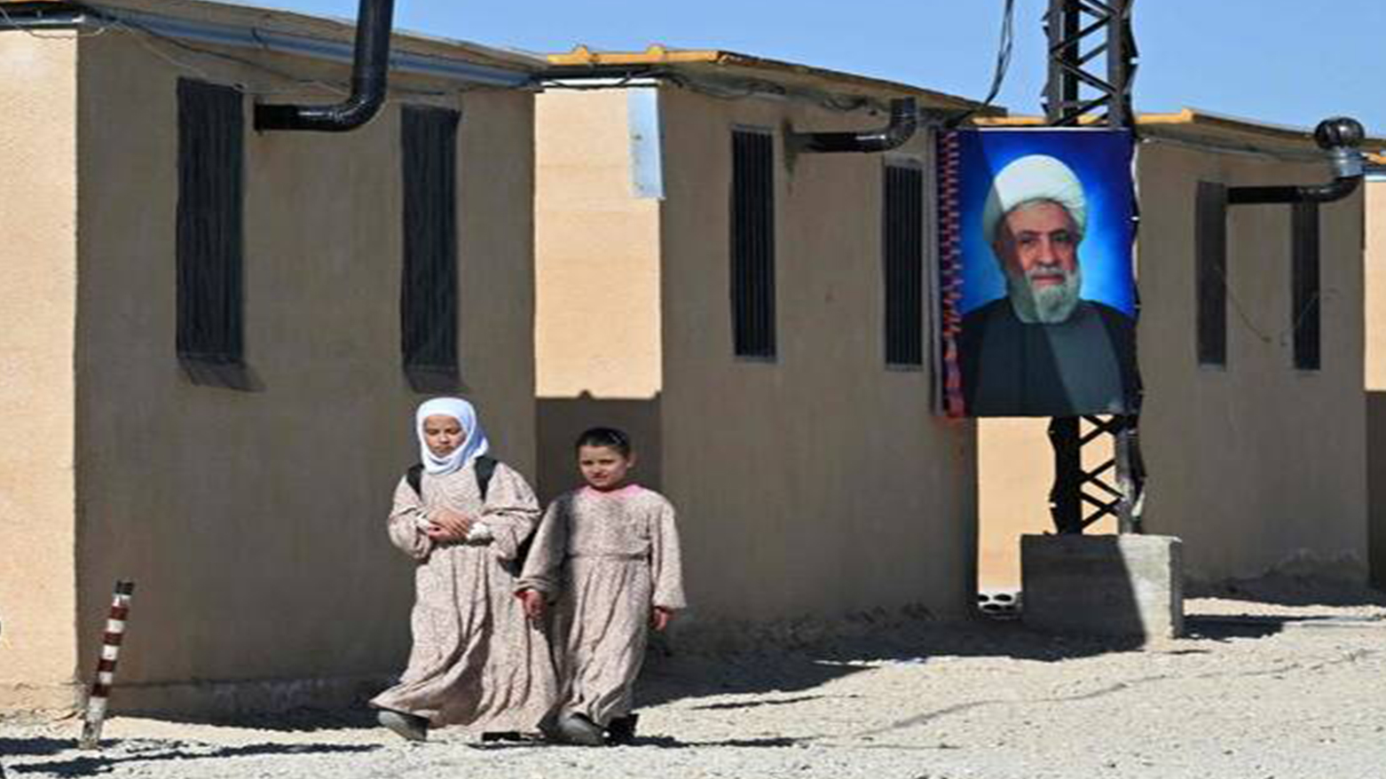 Teenage girls walk past a poster of Hezbollah leader Naim Qassem outside a shelter in Hermel, Lebanon, on Feb. 4, 2026. (AFP)