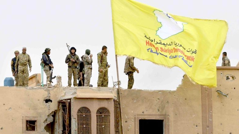 Fighters of the Syrian Democratic Forces stand atop a roof next to their unfurled flag in Baghouz, Deir al-Zor (Photo: AFP)