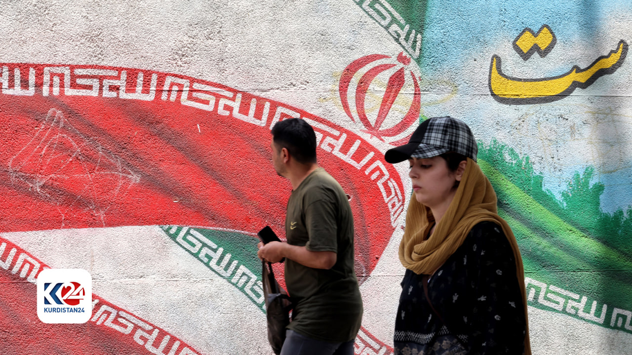 در آستانه دور دوم انتخابات ریاست جمهوری، زن و مردی از کنار یک نقاشی دیواری که پرچم ایران را به تصویر کشیده ‌است، می‌گذرند. تهران، ٢ جولای ٢٠٢٤ – عکس: ای اف پی