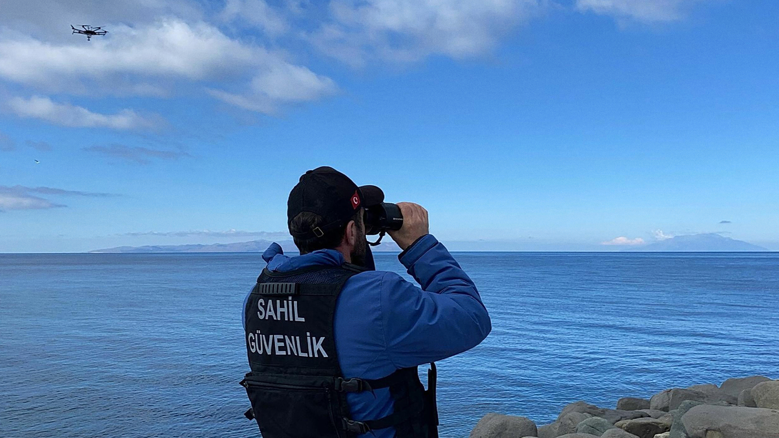 This photograph taken March 15, 2024 in Canakkale shows a coast guard officer looking through binoculars as a drone searches for victims in the Aegean Sea. (Demiroren News Agency/AFP)
