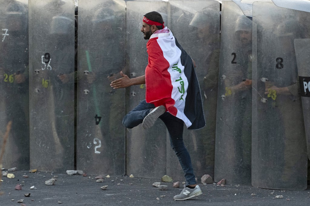 A flag-clad Iraqi protester sprints in front of a wall formed by security officers amid clashes during a rally to mark three years since nationwide demonstrations erupted against endemic corruption, in the southern city of Basra, Oct. 1, 2022. (Photo: Hussein Faleh/AFP)