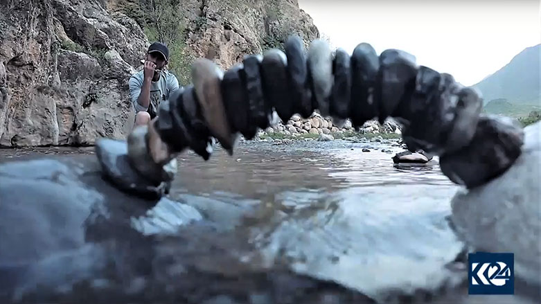 Artist John Safin peacefully plays a harmonica behind one of his natural works of art. (Photo: Kurdistan 24)
