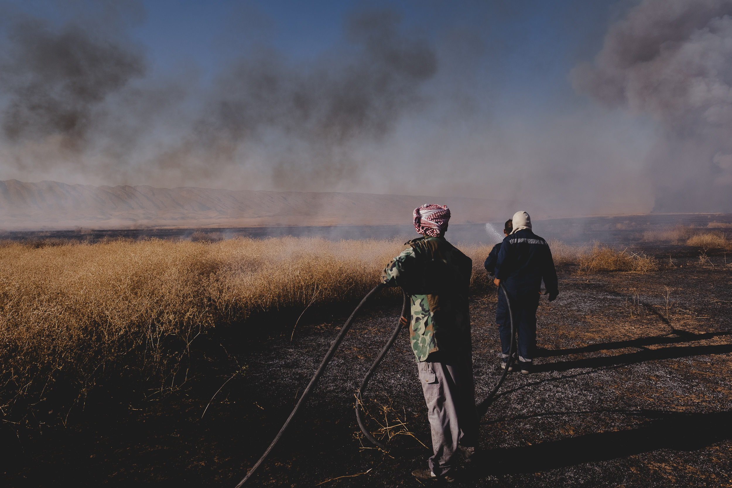 Volunteers from Tel Ezeer carry a hose from a water truck to suppress new patches of fire lit by sparks in the air, with Shingal Mountain to the north. (Photo: Kurdistan 24/Levi Clancy)